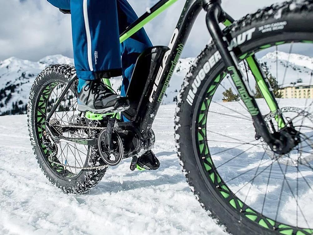 Das Bild zeigt einen Ausschnitt eines Fatbikes mit breiten Reifen, das durch eine verschneite Winterlandschaft gefahren wird. Die Beine des Fahrers in blauer Outdoor-Kleidung sind zu sehen, während die dicke Bereifung des Fahrrads für optimalen Halt im Schnee sorgt. Im Hintergrund sind schneebedeckte Berge und ein Gebäude erkennbar.