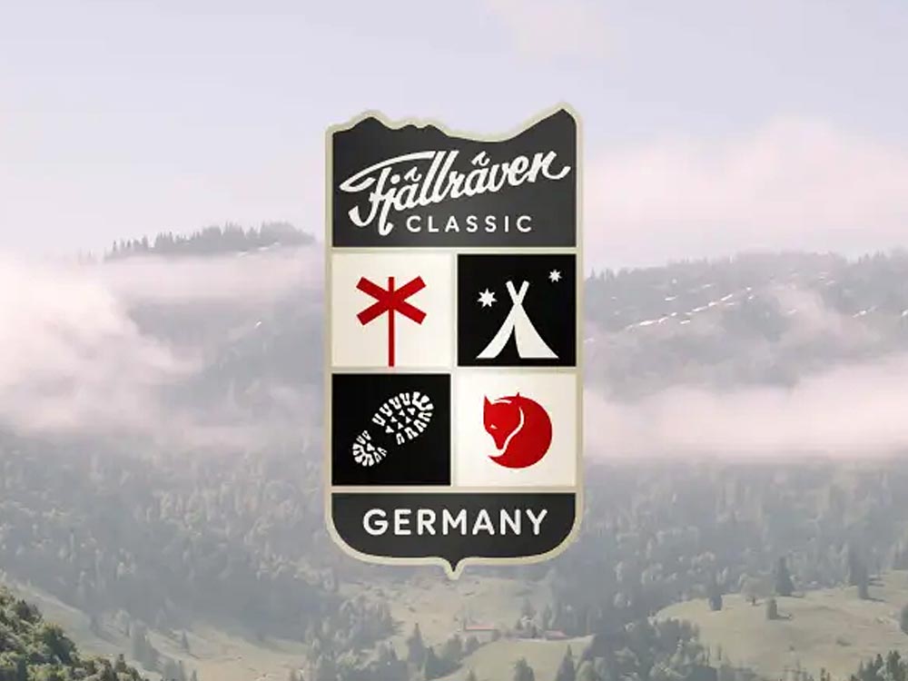 Das Bild zeigt das offizielle Logo der **Fjällräven Classic Deutschland**, ein mehrtägiges Trekking-Event. Das Logo hat die Form eines Schildes mit vier Symbolen: ein rotes Wanderzeichen, ein weißes Zelt mit Sternen darüber, eine schwarze Fußspur und das ikonische rote Fjällräven-Fuchslogo. Darüber steht in geschwungener Schrift **"Fjällräven Classic"**, darunter in Großbuchstaben **"GERMANY"**. Im Hintergrund sieht man eine wunderschöne, nebelverhangene Berglandschaft mit grünen Hügeln und bewaldeten Berghängen. Die Szenerie vermittelt eine friedliche, naturverbundene Atmosphäre, die perfekt zur Outdoor-Philosophie des Events passt.